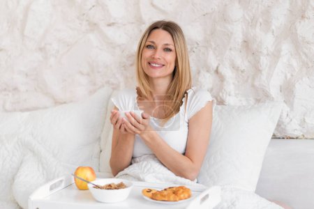 Téléchargez les photos : Joyeux millénaire femme blonde caucasienne en pyjama assis sur le lit, prend le petit déjeuner seul, tenir une tasse de café à l'intérieur de la chambre blanche. Bonjour, dame profiter week-end, temps libre, boisson préférée - en image libre de droit