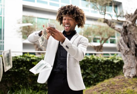 Photo for Overjoyed black middle aged businessman throwing money away showing his wealthiness, standing outdoors in park near office building, free space - Royalty Free Image