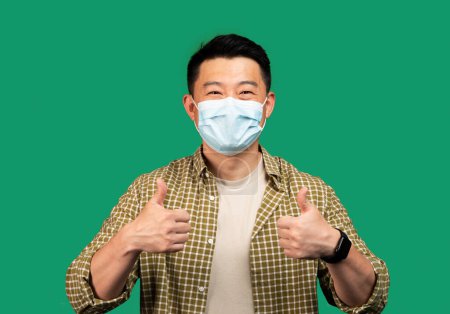 Photo for Asian middle aged man wearing face mask and gesturing thumbs up, approving personal protection on green studio background. Social distance and quarantine, covid-19 prevention concept - Royalty Free Image