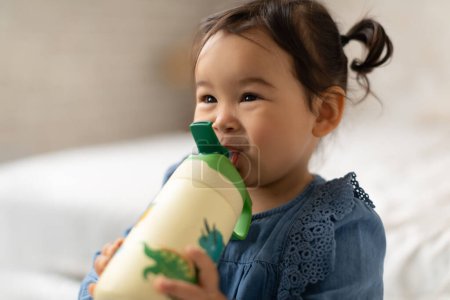 Téléchargez les photos : Hydratation des bébés. Happy Asian Infant Toddler Girl bouteille d'eau potable à la maison. Adorable enfant jouissant d'une hydratation liquide saine à l'intérieur, regardant de l'avant. Concept de nutrition infantile - en image libre de droit