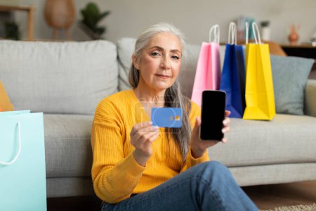 Photo for Serious caucasian old female with a lot of bags sits on floor enjoys shopping with credit card shows phone with blank screen, recommend website in living room interior. Sale, app banking and finance - Royalty Free Image