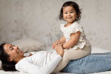Téléchargez les photos : Mignon asiatique bébé fille assis sur gai mère câlins et jouer sourire à la caméra dans moderne chambre à coucher à la maison. Aimer maman coller avec son adorable bébé fille. Congé de maternité - en image libre de droit