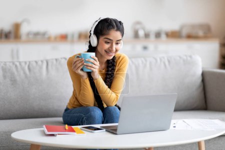 Téléchargez les photos : Femme indienne joyeuse regardant des vidéos sur ordinateur portable et buvant du café à la maison, belle femme orientale heureuse portant des écouteurs sans fil regardant l'écran de l'ordinateur et souriant, espace de copie - en image libre de droit