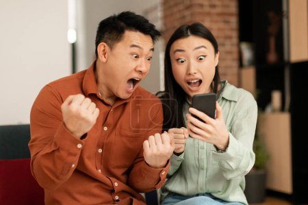 Téléchargez les photos : Couple asiatique émotionnel célébrant le succès, regardant l'écran de téléphone portable et criant dans l'excitation, conjoints acclamant gagner, émotionnellement réagir aux bonnes nouvelles, levant les poings - en image libre de droit