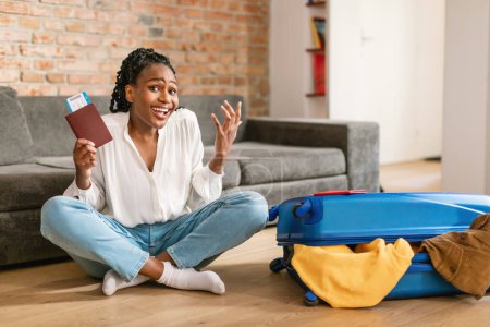 Foto de Mujer afroamericana alegre sosteniendo entradas y agitando puños, celebrando vacaciones, sentada cerca de la maleta empacada en casa. Feliz dama preparándose para el viaje de verano - Imagen libre de derechos