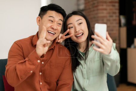 Foto de Emocionada pareja asiática tomando selfie, haciendo gestos de señales de paz y tomando fotos para feliz memoria, redes sociales o entrada de blog. Esposos coreanos tomando fotos en el teléfono inteligente en casa - Imagen libre de derechos