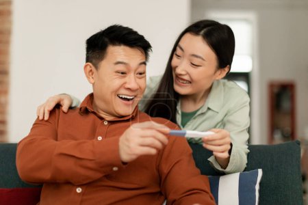 Photo for Happy asian middle aged husband and young wife rejoicing positive pregnancy test at home. Good news, family planning, parenthood and expectation of child indoor - Royalty Free Image