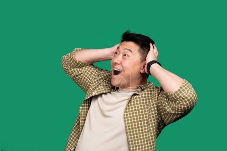 Téléchargez les photos : Choqué asiatique moyen âge homme ouverture bouche et toucher la tête, surpris mâle debout sur fond de studio vert, émotionnellement réagir aux nouvelles - en image libre de droit