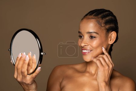Photo for Beauty care concept. Attractive black middle aged woman holding magnifying mirror, looking at reflection and enjoying her perfect skin, posing over brown background - Royalty Free Image