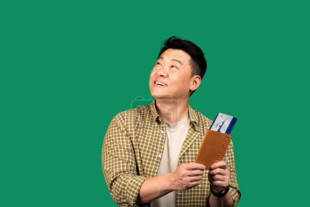 Téléchargez les photos : Portrait d'un homme asiatique d'âge moyen tenant un passeport avec des billets d'avion, regardant de côté l'espace de copie sur fond de studio vert. Tourisme outre-mer, concept de vacances à l'étranger - en image libre de droit