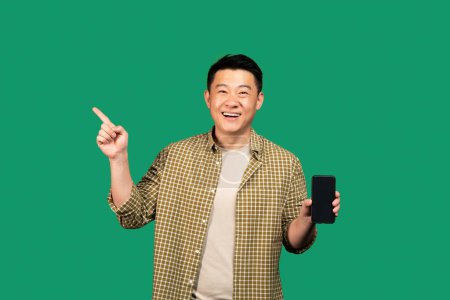 Foto de Feliz asiático de mediana edad hombre mostrando el teléfono celular con la pantalla vacía y apuntando a un lado en el espacio libre, recomendando aplicación móvil o sitio web en fondo verde, maqueta - Imagen libre de derechos