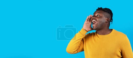 Téléchargez les photos : Jeune homme noir excité avec la main près de la bouche faisant l'annonce, homme afro-américain joyeux partageant des nouvelles ou des informations tout en restant isolé sur fond bleu, Panorama, Copier l'espace - en image libre de droit