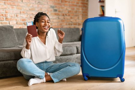 Photo for Excited african american female tourist with passport and flight tickets clenching fists while sitting by big packed suitcase at home, ready for trip - Royalty Free Image