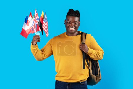 Téléchargez les photos : Portrait d'un étudiant noir souriant avec sac à dos tenant des drapeaux internationaux tout en se tenant sur fond bleu en studio, homme afro-américain heureux bénéficiant d'une éducation à l'étranger, espace de copie - en image libre de droit