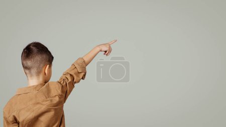 Foto de Caucásico niño de 6 años de edad en casual señalar el dedo en el espacio vacío aislado sobre fondo gris estudio, espalda, panorama. Estilo de vida infancia, educación, estudio, anuncio y oferta - Imagen libre de derechos