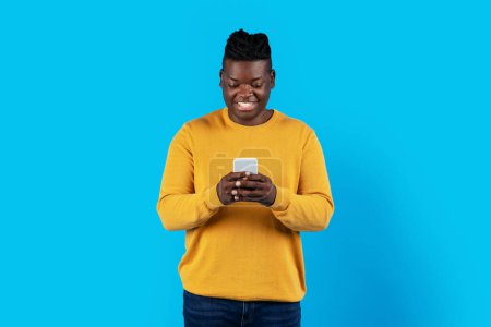 Photo for Portrait Of Young Smiling Black Guy Using Smartphone For Online Communication, Happy African American Male Texting On Cellphone With Friends While Standing Isolated Over Blue Background, Copy Space - Royalty Free Image