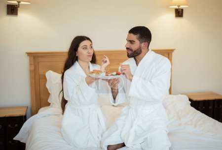 Foto de Los cónyuges que comen el desayuno en la cama con albornoces blancos en el interior. Pareja casada disfrutando de una cita romántica en el hotel degustando sándwiches por la mañana. Nutrición saludable en vacaciones - Imagen libre de derechos