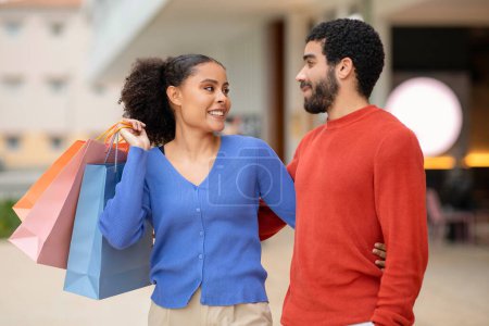 Photo for Shopping With Boyfriend. Happy Multiracial Couple Holding Paper Shopper Bags Spending Time In Modern Mall Indoors On Weekend, Smiling To Each Other. Commerce And Sales Offer - Royalty Free Image