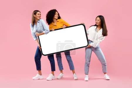 Photo for Excited diverse female friends holding huge smartphone with empty screen on pink studio background, mockup for mobile app or new website. Cell phone display template, free space, full body length - Royalty Free Image