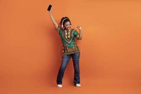 Téléchargez les photos : Positif cool heureuse femme noire millénaire attrayante en chemise africaine nationale bénéficiant d'une belle application de musique, écouter de la musique et de la danse, en utilisant des écouteurs stéréo sans fil, smartphone, fond orange - en image libre de droit
