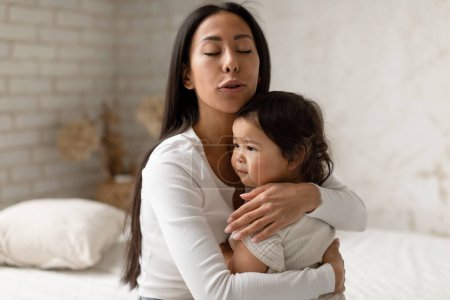 Téléchargez les photos : Caring Korean Mom Calmer pleurer bébé fille étreignant bébé fille assise sur le lit dans la chambre moderne à la maison. Mère apaisant son tout-petit bouleversé. Concept de maternité et de garde d'enfants - en image libre de droit