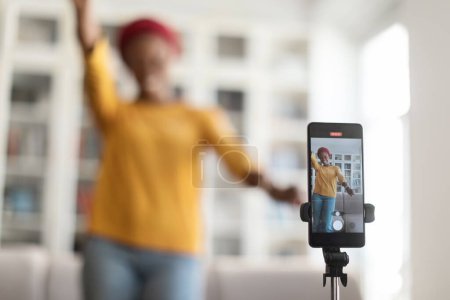 Photo for Selective focus on modern smartphone set on tripod recording female influencer cheerful young black woman in casual wearing african turban dancing at home, blurred background. Blogging concept - Royalty Free Image