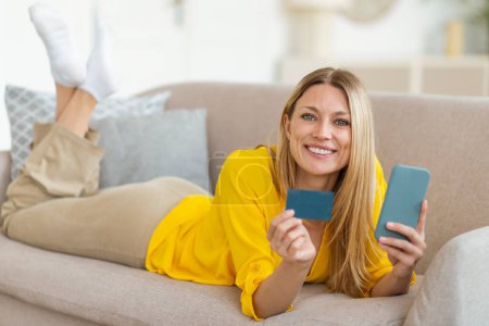 Photo for Cashback for shopaholic. Smiling millennial caucasian blonde female in yellow clothes with credit card and smartphone shopping online in living room interior. Sale, financial account check, money app - Royalty Free Image