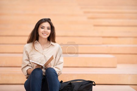 Téléchargez les photos : Souriant millénaire mixte dame brune étudiante siège et lit livre dans les escaliers du campus à l'université, en plein air. Rompre avec l'étude, les connaissances, l'éducation, le mode de vie dans la publicité et l'offre collégiale - en image libre de droit