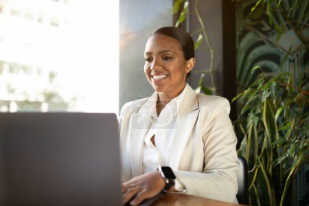 Foto de Positiva mujer de negocios afroamericana milenaria en traje blanco que trabaja en la computadora portátil en la oficina de la cafetería con el interior de las plantas verdes. Estilo de vida empresarial, nómada digital y gadget - Imagen libre de derechos