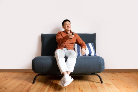 Foto de Feliz asiático de mediana edad hombre viendo televisión celebración mando a distancia, sentado en sofá, divertirse en blanco fondo de la pared. Programación de televisión, concepto de ocio fin de semana - Imagen libre de derechos