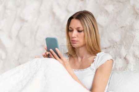 Photo for Serious sad millennial caucasian blonde female in domestic clothes sits on bed, reads message on phone in white bedroom interior. Reaction to bad news, app and social networks, problems and stress - Royalty Free Image