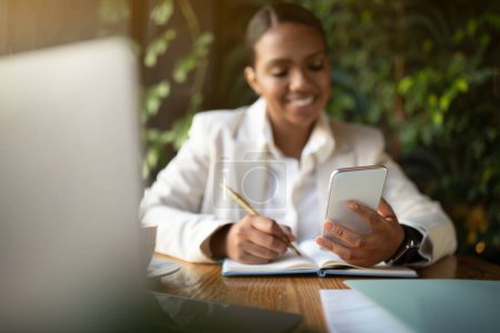 Foto de Feliz millennial mujer de negocios afroamericana en traje blanco mensaje de lectura en el teléfono inteligente, utiliza el ordenador portátil en eco café con el interior de las plantas verdes. Aplicación para negocios, trabajar con gadget de forma remota - Imagen libre de derechos