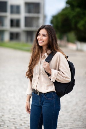 Téléchargez les photos : Souriant jolie étudiante brune caucasienne millénaire décontractée avec sac à dos regarde l'espace vide sur le campus à l'université, en plein air, vertical. Etude, connaissance et éducation - en image libre de droit