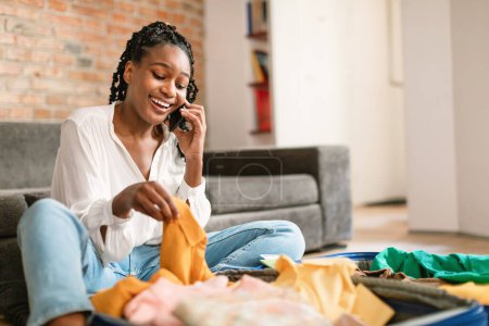 Téléchargez les photos : Femme noire excitée se préparant pour les vacances, parler sur smartphone et valise, réserver un voyage, faire une réservation d'hôtel au téléphone à la maison - en image libre de droit