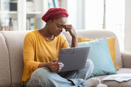 Téléchargez les photos : Épuisé contrarié jeune femme noire en tenue décontractée turban rouge assis sur le canapé, en utilisant l'ordinateur à la maison, toucher la tête, travailler en ligne, se sentir fatigué, copier l'espace. Concept d'épuisement professionnel - en image libre de droit