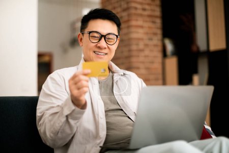 Photo for Positive middle aged asian man sitting on sofa, using modern laptop and credit card at home, shopping or banking on Internet, making online order, copy space - Royalty Free Image