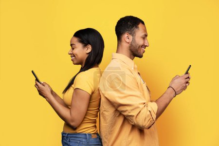 Photo for Glad millennial african american guy and female typing on smartphone, surfing in internet, isolated on yellow studio background, profile. Chat in social networks, device for communication - Royalty Free Image
