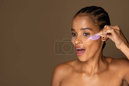 Téléchargez les photos : Femme afro-américaine excitée décollant les patchs hydrogel, debout isolé sur fond brun, espace libre. Spa à la maison, soins quotidiens, cosmétiques pour soins de beauté - en image libre de droit