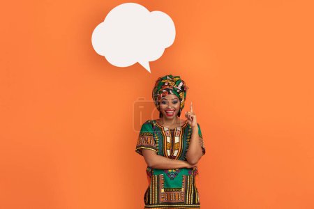 Foto de Emocionado joven mujer negra feliz en traje africano con burbuja conversación en blanco sobre su cabeza en fondo de estudio naranja, apuntando con el dedo hacia arriba, eureka, espacio para el diseño, señora tiene buenas ideas - Imagen libre de derechos