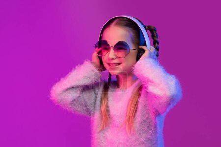 Téléchargez les photos : Portrait de mignonne jolie jolie petite fille blonde d'âge scolaire en lunettes de soleil utilisant des écouteurs sans fil modernes, enfant heureux écoutant de la musique, s'amusant sur fond de studio rose dans la lumière néon - en image libre de droit