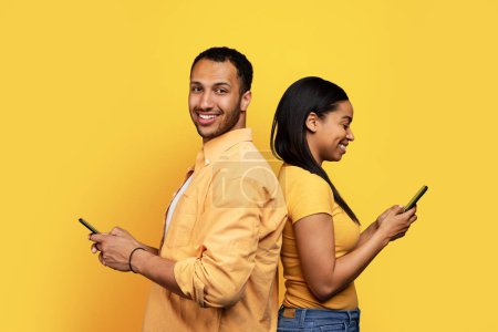 Foto de Glad millennial afroamericano masculino y femenino escribiendo en el teléfono inteligente, disfrutar de charlar juntos aislados en el fondo del estudio amarillo. Redes sociales para romántico, dispositivo para la comunicación - Imagen libre de derechos