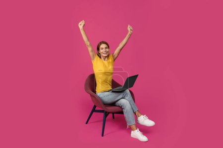Foto de Mujer europea adulta satisfecha sentada en silla con portátil, levantando las manos, celebrando el éxito y la victoria, aislada sobre fondo púrpura, estudio. Gran oferta y anuncio, venta, gadget para chat y blog - Imagen libre de derechos