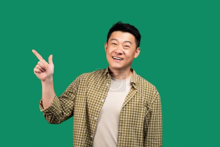 Téléchargez les photos : Excité asiatique homme d'âge moyen pointant du doigt à l'espace libre, offre de publicité sur fond de studio vert et souriant. Regardez là concept - en image libre de droit