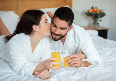 Téléchargez les photos : Happy Wife Embrasser les maris joue Boire du jus d'orange se détendre dans la chambre d'hôtel en vacances. Couple multiethnique jouissant d'une lune de miel romantique ensemble à l'intérieur. Offre Spa Resort - en image libre de droit