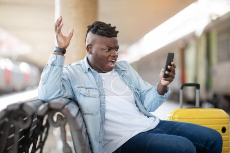 Téléchargez les photos : Stressé Black Guy lecture mauvais message sur Smartphone tout en étant assis à la gare, Furieux Afro-Américain Homme souffrant de problèmes avec téléphone portable en attendant le train sur la plate-forme - en image libre de droit
