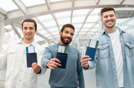 Téléchargez les photos : Offre de voyage. Trois voyageurs masculins joyeux montrant leurs passeports avec des passes d'embarquement à la caméra posant debout avec des valises dans l'aéroport moderne à l'intérieur. Focus sélectif - en image libre de droit