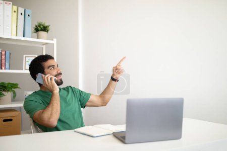 Téléchargez les photos : Homme arabe millénaire joyeux avec des appels de barbe par téléphone, travaille avec ordinateur portable sur le lieu de travail, pointe du doigt l'espace vide à l'intérieur de la salle de bureau. Affaires, communication client à distance à la maison - en image libre de droit