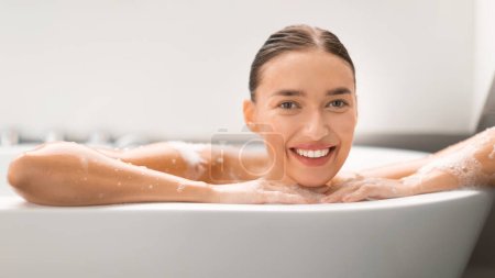 Téléchargez les photos : Superbe femme prenant bain posant dans la baignoire souriant à la caméra dans la salle de bain moderne à la maison. Lady Bain Soins pour le corps recouvert de savon Sud. Concept de routine beauté. Panorama - en image libre de droit