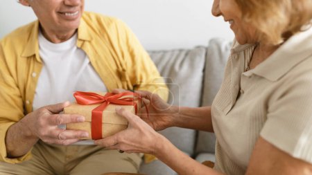 Téléchargez les photos : Senior homme donnant coffret cadeau à femme heureuse, la félicitant avec anniversaire ou anniversaire, assis sur le canapé à la maison, recadré. Conjoints âgés célébrant des vacances en famille ensemble - en image libre de droit