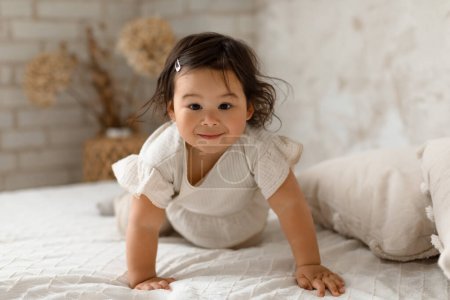 Téléchargez les photos : Japonais tout-petit bébé fille rampant en regardant la caméra sur le lit dans la chambre moderne à l'intérieur. Mignon bébé posant parmi les oreillers explorant sa chambre à la maison. Happy Babyhood Concept - en image libre de droit